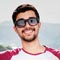 A person with short dark hair and a beard is smiling, wearing dark sunglasses and a red and white shirt, possibly enjoying a break from working on bookkeeping software. The background appears to be an outdoor setting with trees visible in the distance.